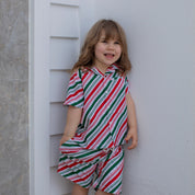 A young girl with blonde hair stands barefoot against a white wall, wearing a matching short-sleeve collared shirt and shorts set with diagonal red, green, and white stripes.
