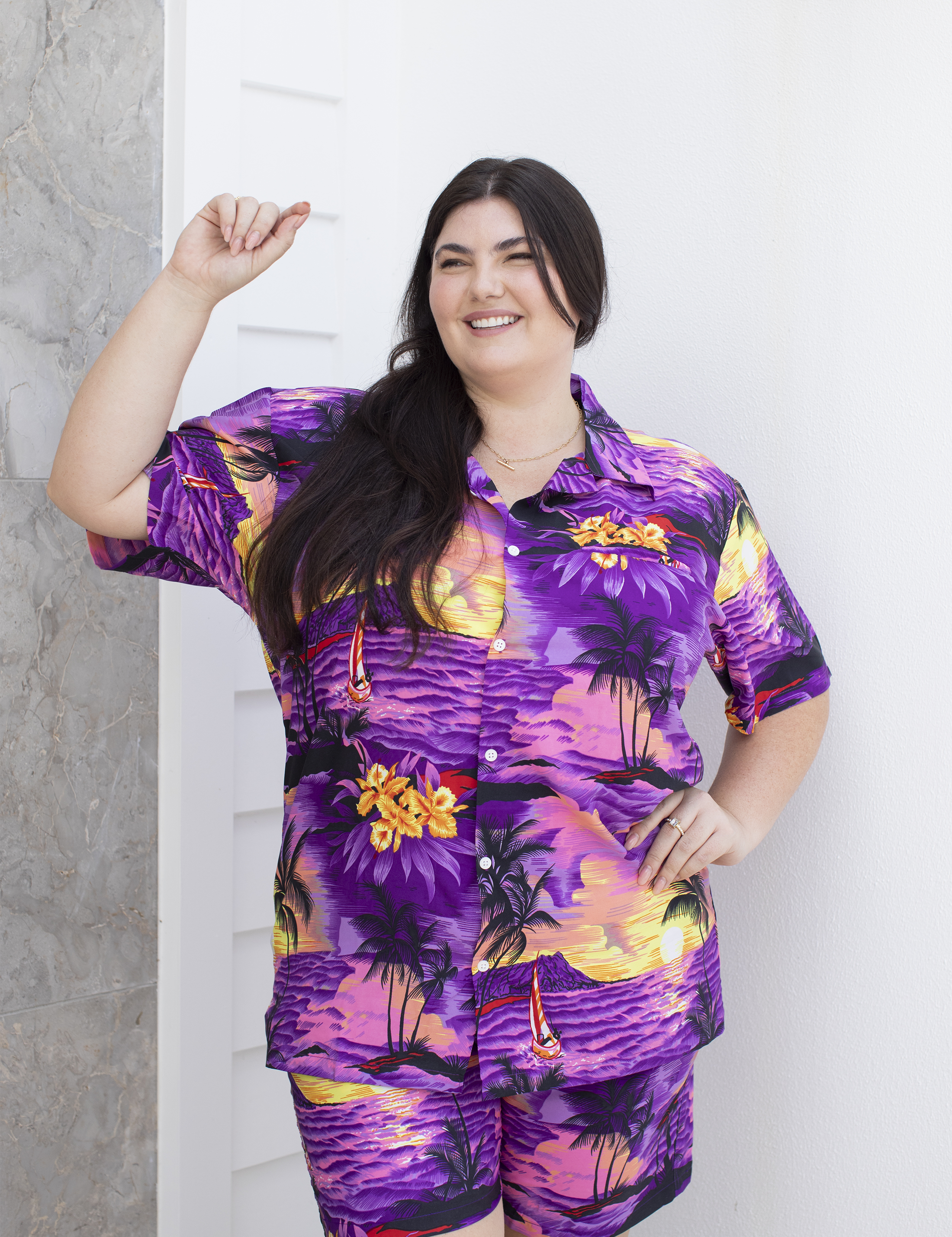 Adult female model wearing a purple Hawaiian shirt and shorts with a tropical design, posing with one hand extended.