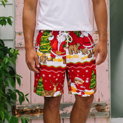 A man stands on a white porch wearing a white crewneck t-shirt and red Christmas-themed swim trunks with a Santa Claus and reindeer print. The background features a pink wooden door, white siding, and green foliage with pink flowers.