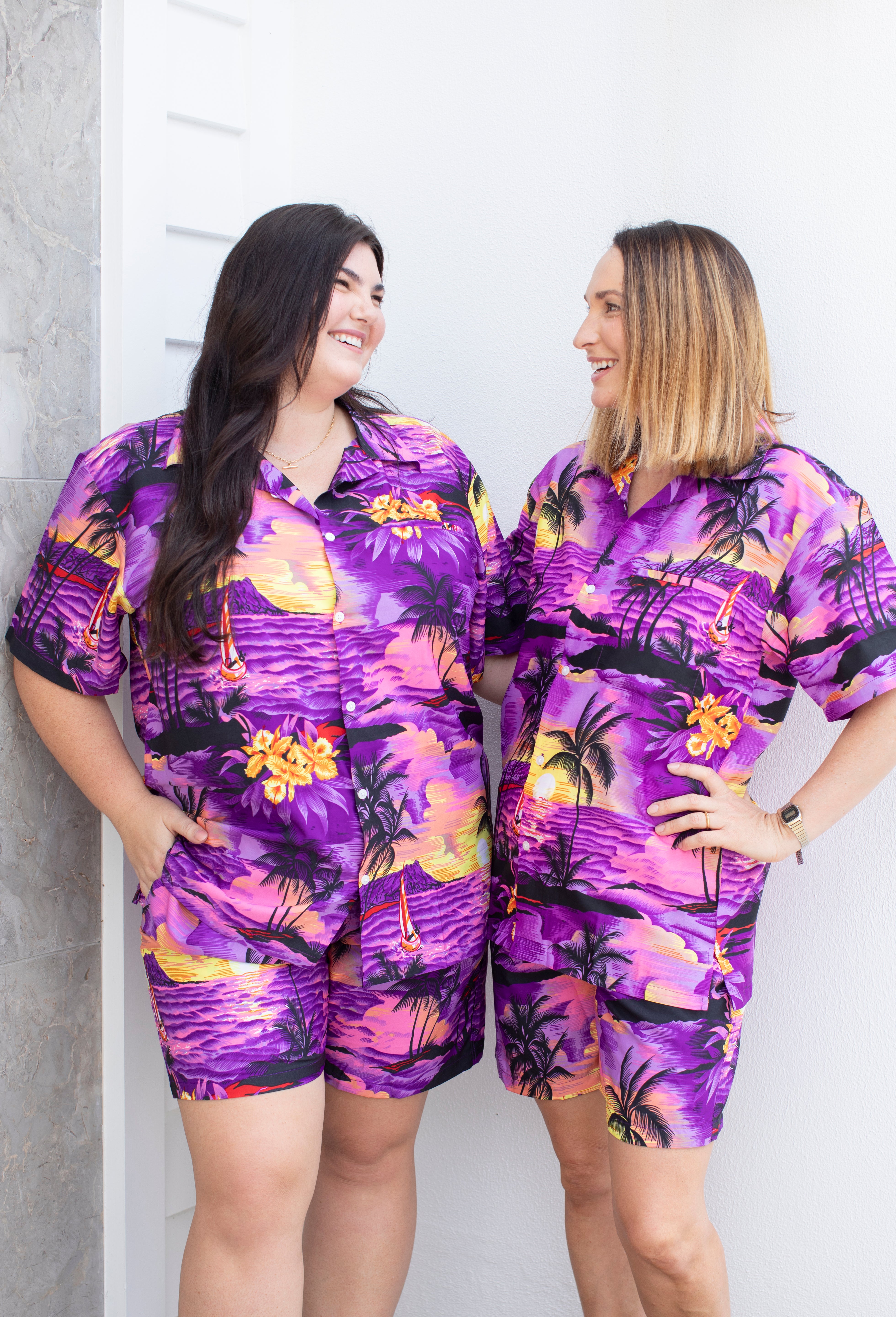 Two women wearing matching purple tropical outfits standing against a white wall.