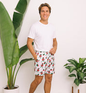 A man wearing a white t-shirt and white shorts with a red and green Santa Claus pattern, standing in a brightly lit room with potted plants.