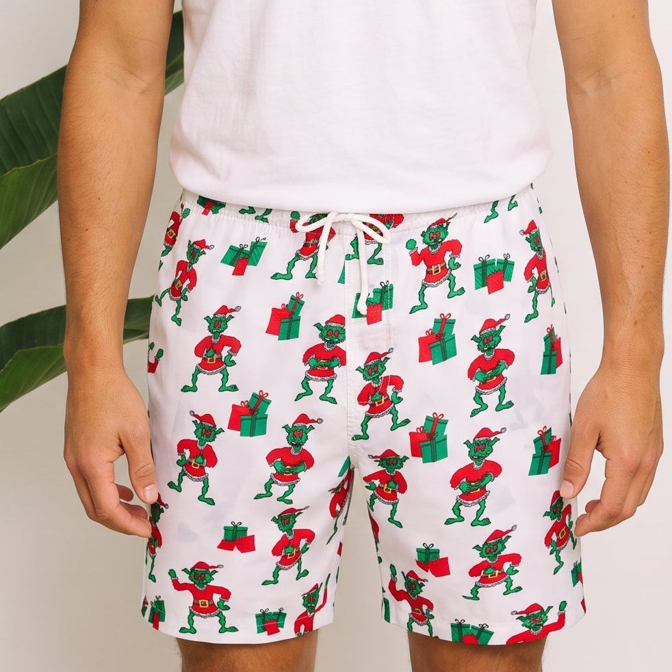A man wearing a white crewneck t-shirt and white drawstring shorts with a green and red Santa Claus pattern, standing in front of a white wall with tropical plants.