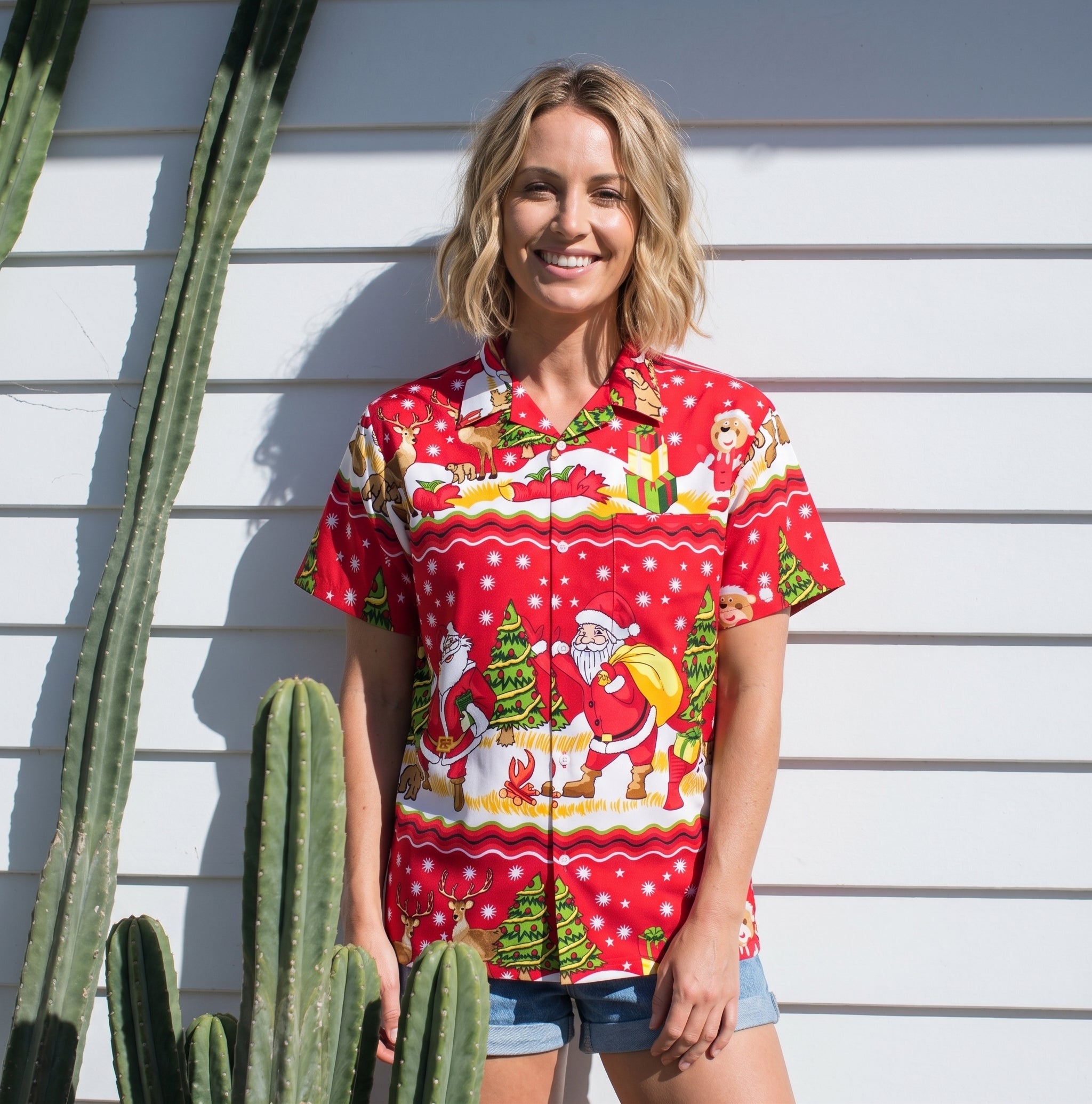 A woman stands outdoors against a white siding wall wearing a bright red short-sleeve button-up shirt with a festive Christmas print featuring Santa Claus, a dog, trees, and snowflakes. She is also wearing denim shorts, with tall cacti visible on the left side of the frame.