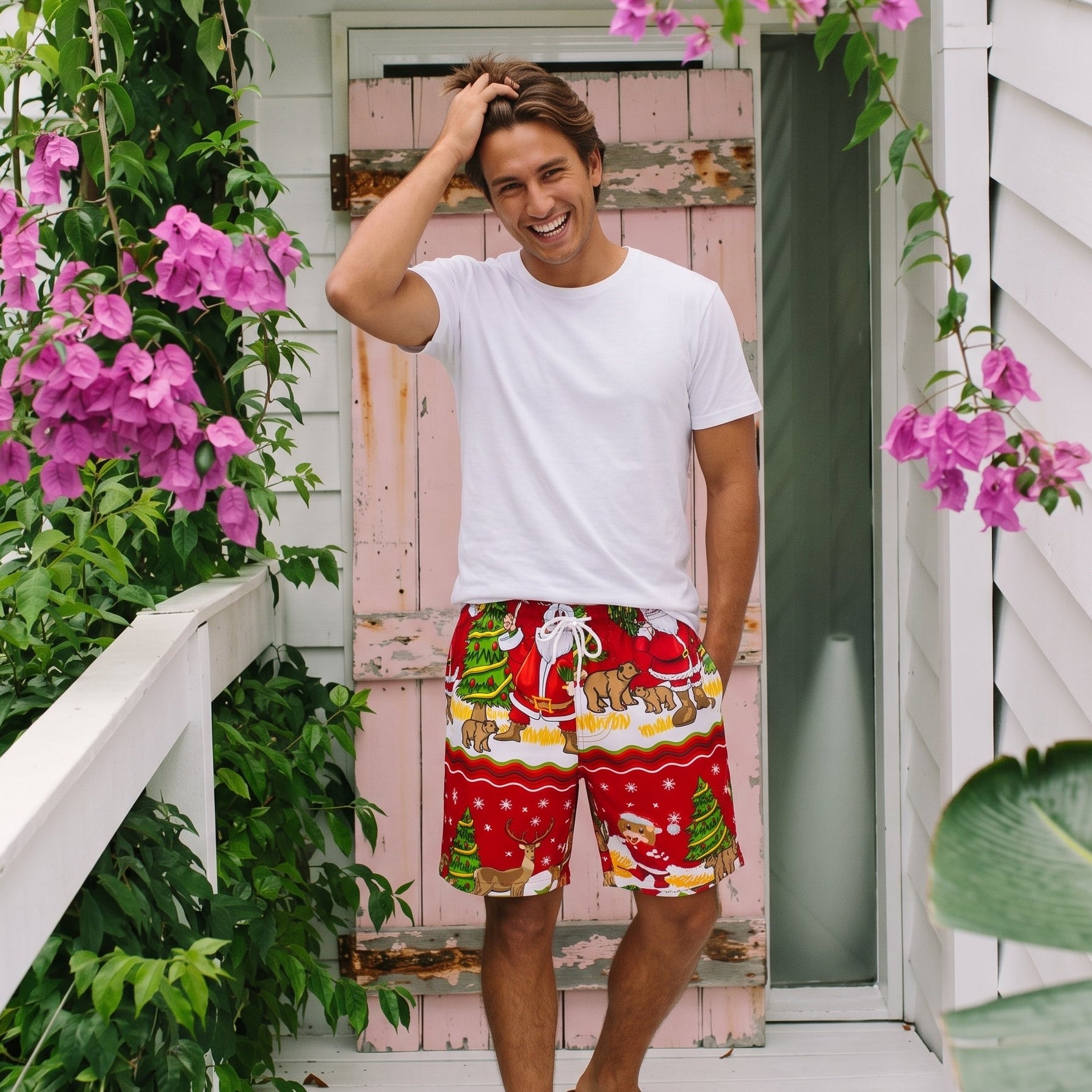 A man stands on a white porch wearing a white t-shirt and red Christmas-themed shorts with Santa, reindeer, and trees, surrounded by green plants and pink flowers.