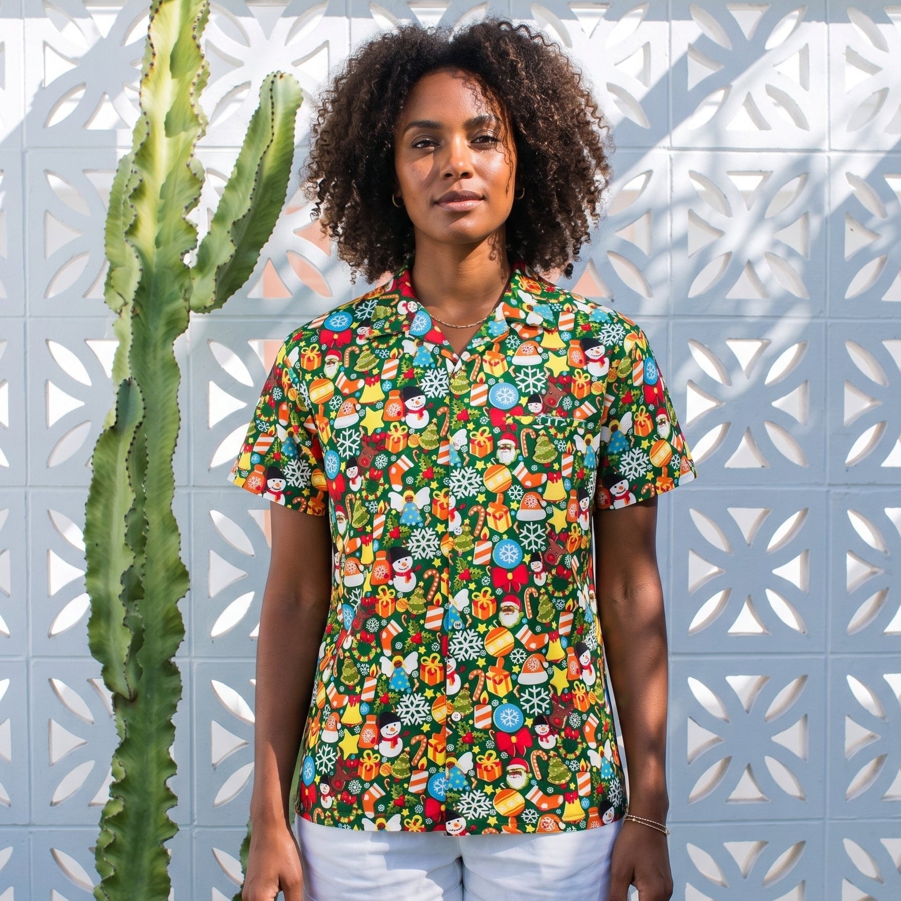 A woman stands outdoors wearing a short-sleeve button-up scrub top with a dense multicolor floral and botanical print, paired with white shorts. She is positioned next to a tall green cactus in front of a white geometric patterned wall.