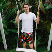 A man stands on a white rope swing in a lush green outdoor setting, wearing a white t-shirt and black swim trunks with a colorful Christmas-themed print of Santa Claus, gingerbread men, and snowballs.