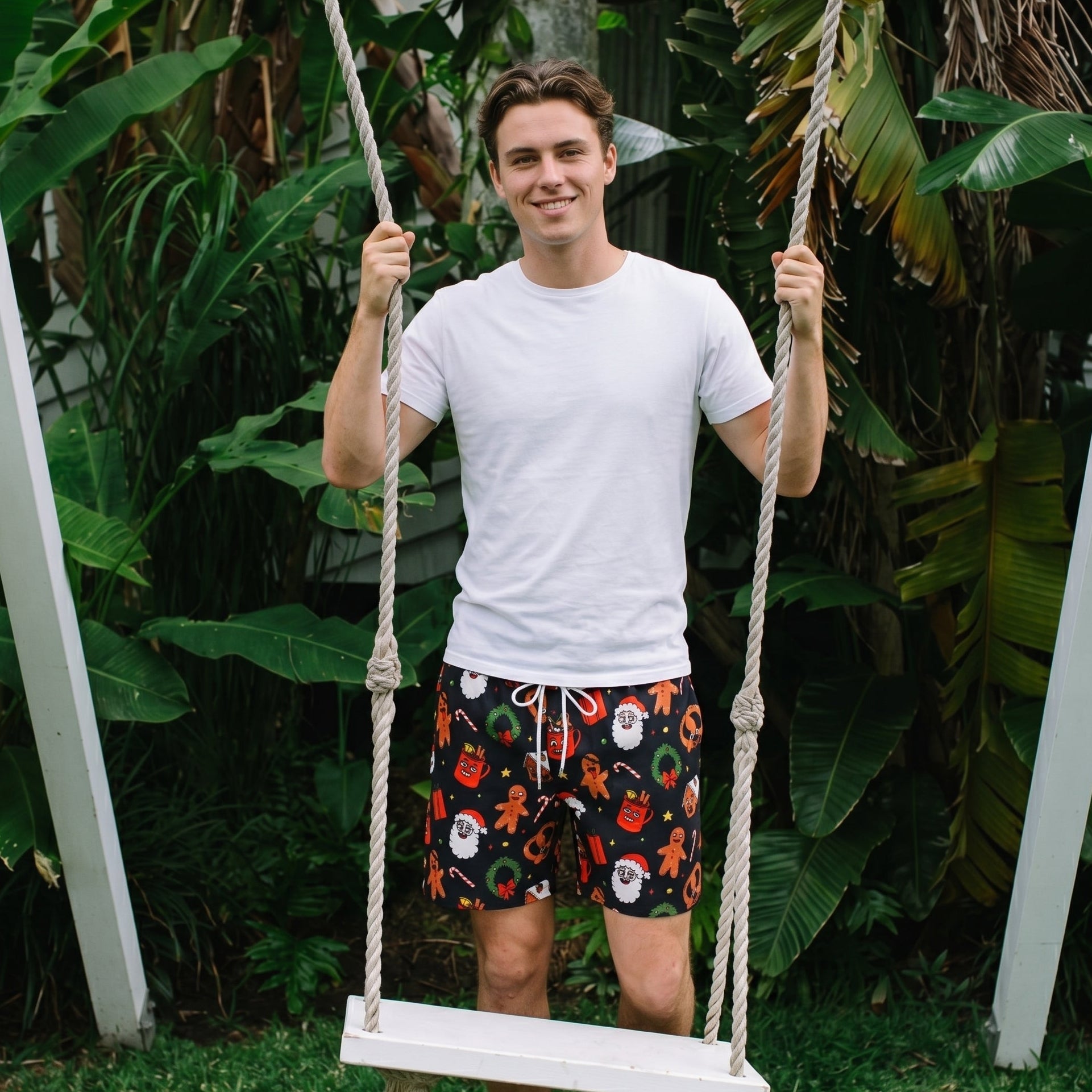 A man stands on a white rope swing in a lush green outdoor setting, wearing a white t-shirt and black swim trunks with a colorful Christmas-themed print of Santa Claus, gingerbread men, and snowballs.