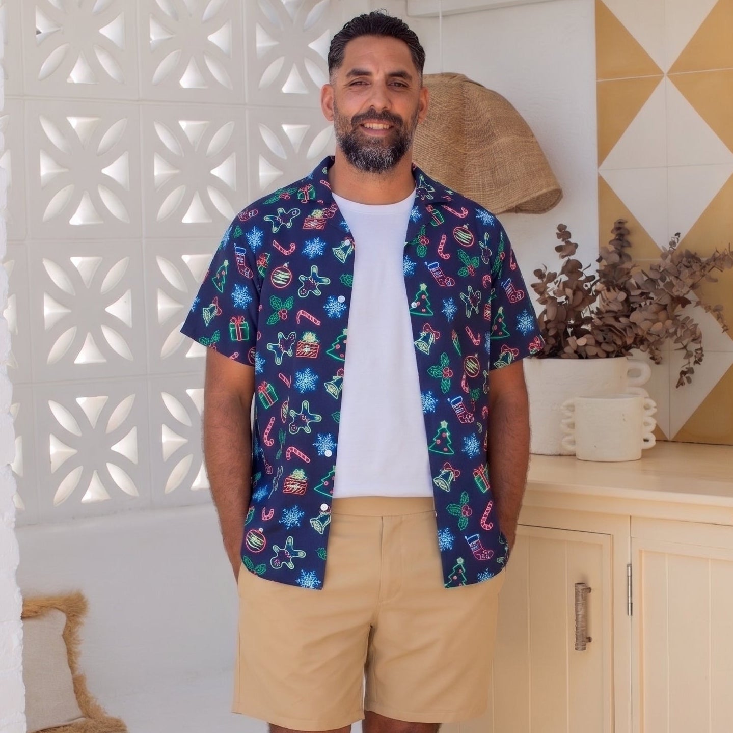 A man stands barefoot in a bright, modern room wearing a short-sleeve button-up shirt with a colorful tropical print, a white t-shirt, and khaki shorts.