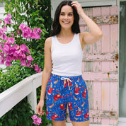 A woman stands outdoors wearing blue patterned shorts with a white tank top. The shorts feature a colorful print of Santa Claus, snow globes, and a white drawstring.