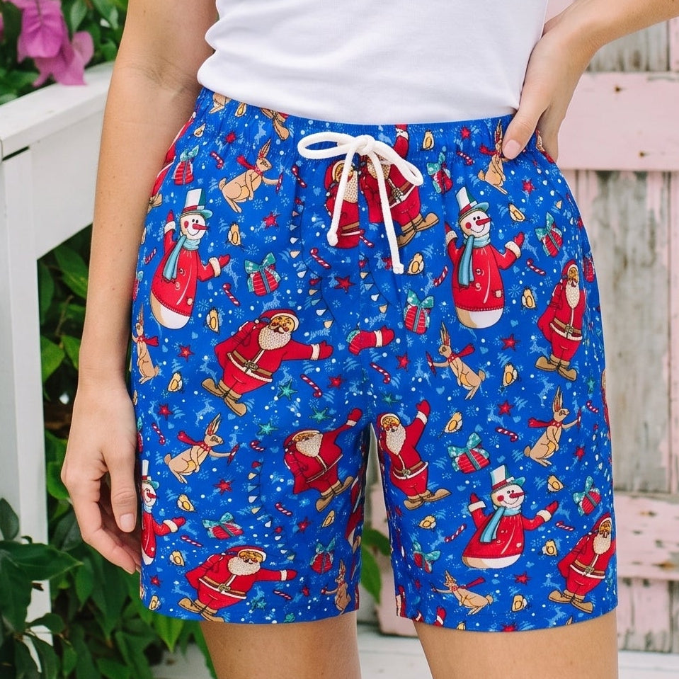 A woman modeling blue holiday-themed shorts with a Santa Claus print, paired with a white tank top, standing outdoors in front of a pink wooden door and green foliage with pink flowers.