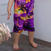 A barefoot child stands on a wooden deck, showing only their lower half and wearing Sunset Purple Kids Shorts—a vibrant choice paired with a matching tropical shirt featuring palm trees, flowers, and a sunset pattern for festive family gatherings.
