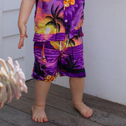 A barefoot child stands on a wooden floor, wearing Sunset Purple Kids Shorts paired with a vibrant tropical outfit featuring palm trees and sunset designs—ideal for festive family gatherings. Only the lower half of the child's body is visible.