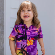 A smiling young child with long light brown hair showcases the Sunset Purple Kids Shirt, which features a vibrant design of palm trees and flowers, while standing outdoors in radiant sunlight.