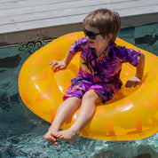 A young boy in a Sunset Purple Kids Shirt, featuring a bright design, sits on a yellow inflatable float in a pool, smiling as his feet dangle in the water beside a wooden deck.