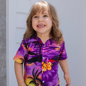 A smiling young child with long light brown hair wears the Sunset Purple Kids Shirt, featuring a bright design, while standing outside by a light-colored wall.