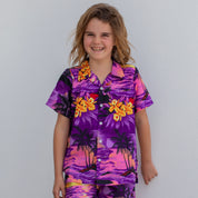 A smiling child with wavy hair stands against a white wall, wearing the Sunset Purple Kids Shirt adorned with yellow flowers and palm trees, paired with matching shorts.