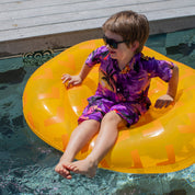 A young boy in sunglasses and a Sunset Purple Kids Shirt with a vibrant print relaxes on a yellow pool float, smiling with his feet in the water by a wooden deck.
