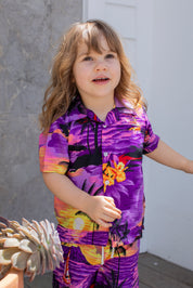 A young child with long, wavy hair wears the Sunset Purple Kids Hawaiian Shirt and Shorts Set, standing outdoors by a gray wall and a potted succulent.