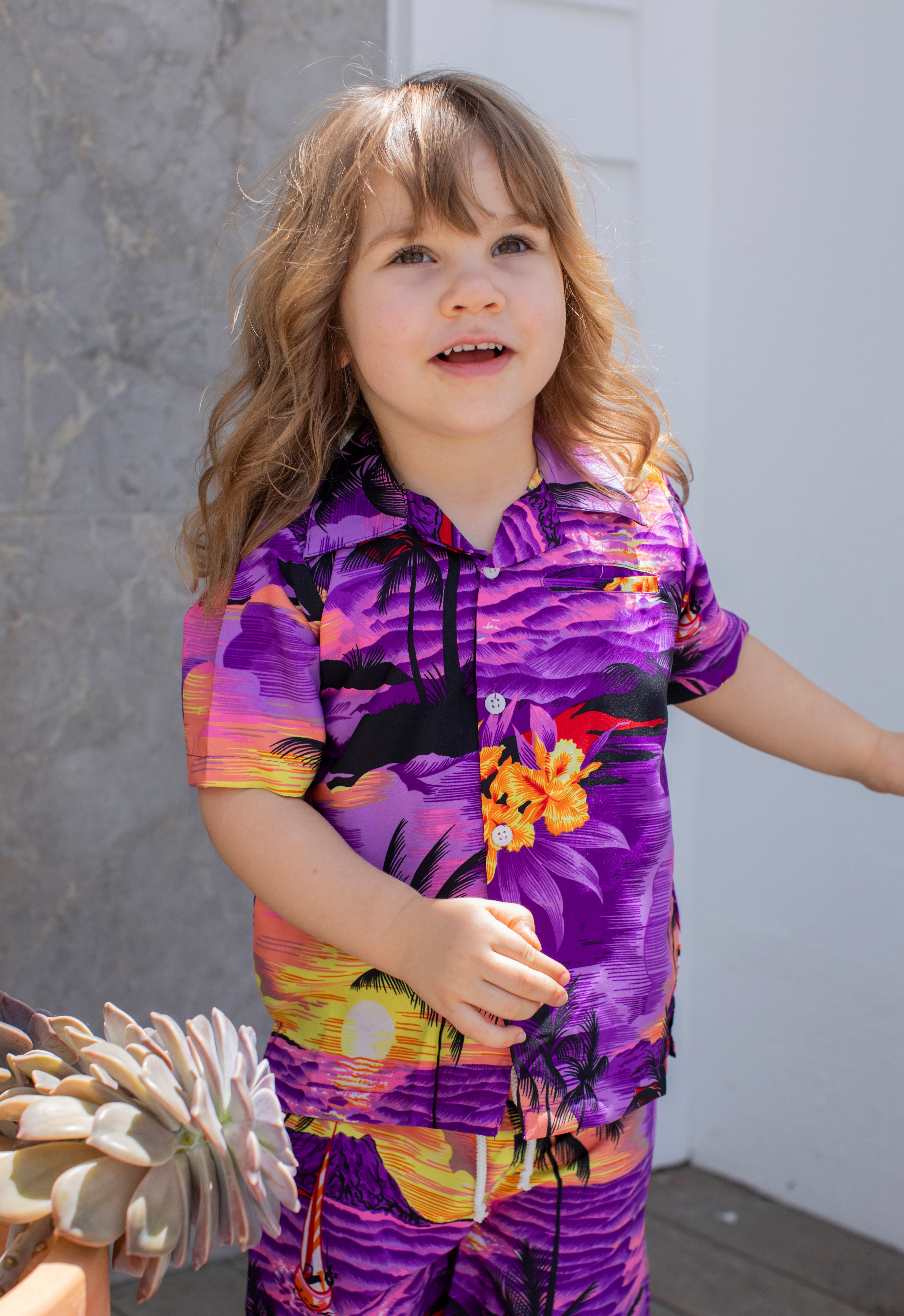 A young child with long, wavy hair wears the Sunset Purple Kids Hawaiian Shirt and Shorts Set, standing outdoors by a gray wall and a potted succulent.