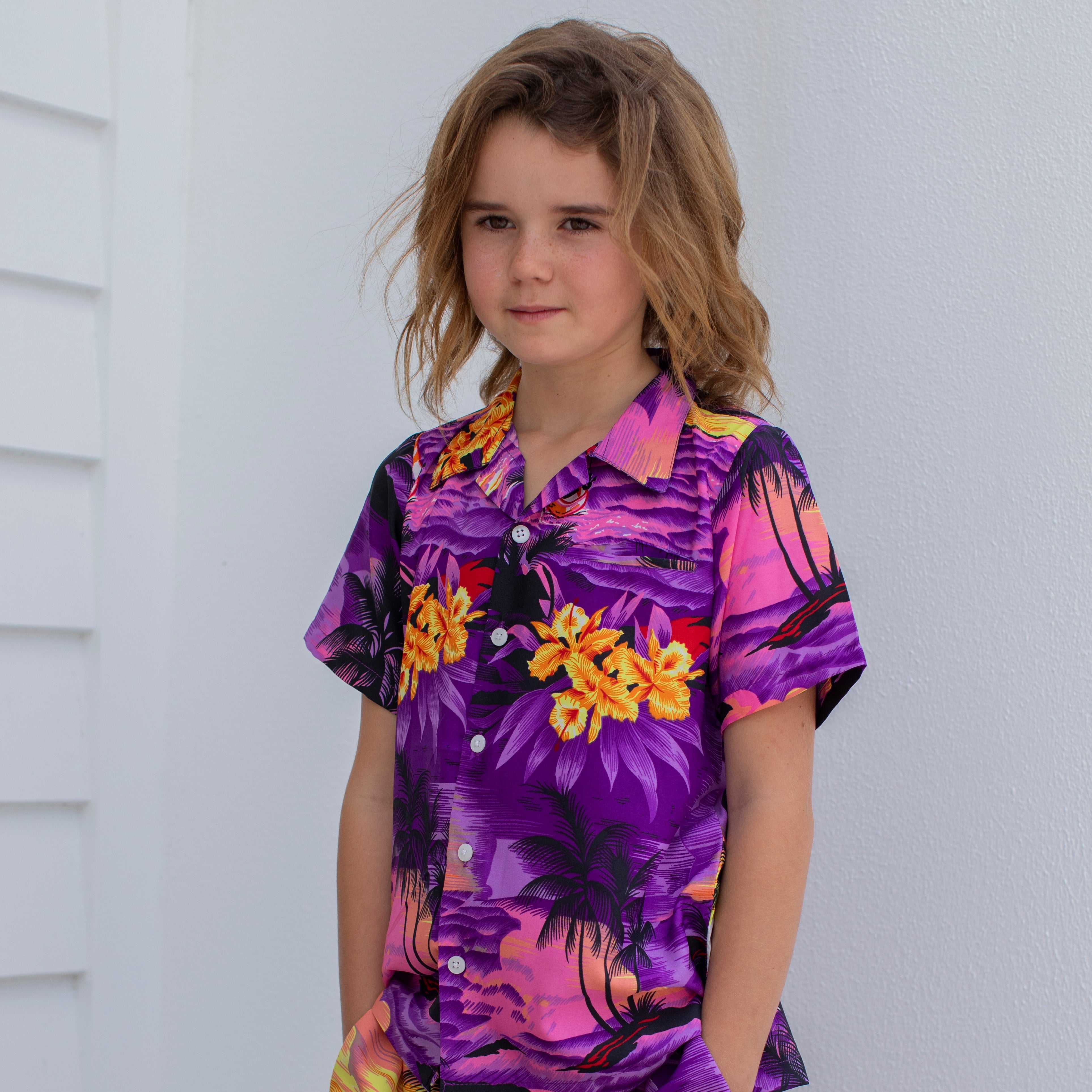 A young child with long brown hair wears the Sunset Purple Kids Hawaiian Shirt and Shorts Set, featuring a vibrant tropical print, and stands in front of a white wall looking slightly to the side with a neutral expression.