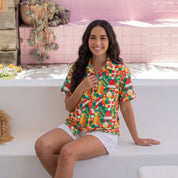 A woman sits on a white outdoor bench wearing a colorful short-sleeve button-up shirt with a bold tropical fruit and candy print, paired with white shorts and beige slide sandals, set against a pink tiled wall background.