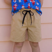 A young boy with blonde hair stands against a peach-colored house siding, wearing a blue short-sleeve button-up shirt with a Santa Claus pattern, tan shorts, and white sneakers.